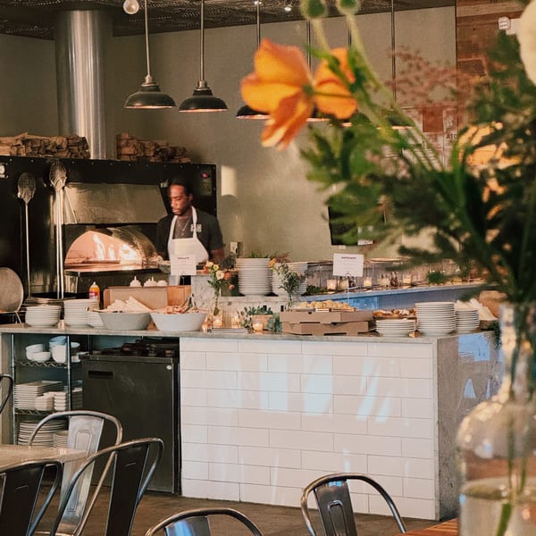 A chef stands by a wood-fired oven in a restaurant with industrial and rustic decor.