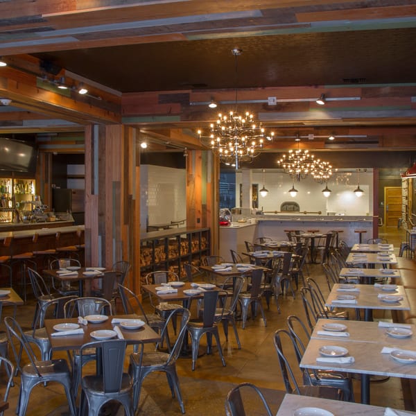 Spacious restaurant interior with a bar, dining tables, and an open kitchen with a pizza oven.