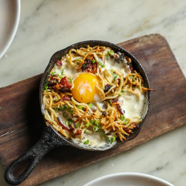 Skillet of creamy noodles with bacon, green onions, and a sunny-side-up egg on a wooden board.