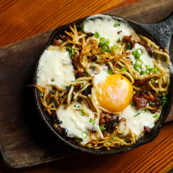 Skillet of crispy noodles, bacon, melted cheese, and a sunny-side-up egg on a wooden board.