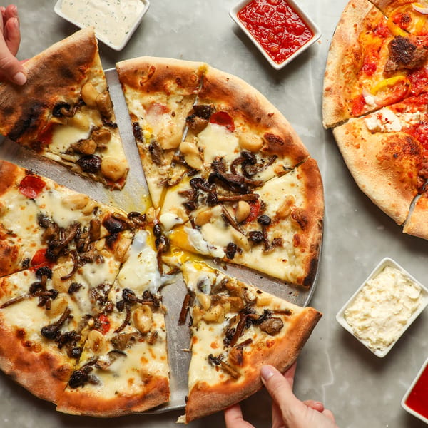 Two hands grab slices from a mushroom and egg pizza, with another pizza and dipping sauces nearby.