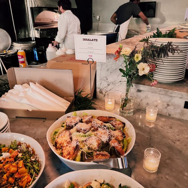Restaurant kitchen with chefs, a pizza oven, and a buffet of salads and stacked plates.