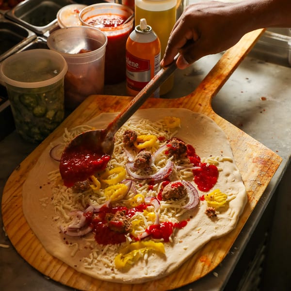 A chef's hand spoons red sauce onto a pizza with cheese, onions, and sausage on a wooden peel.